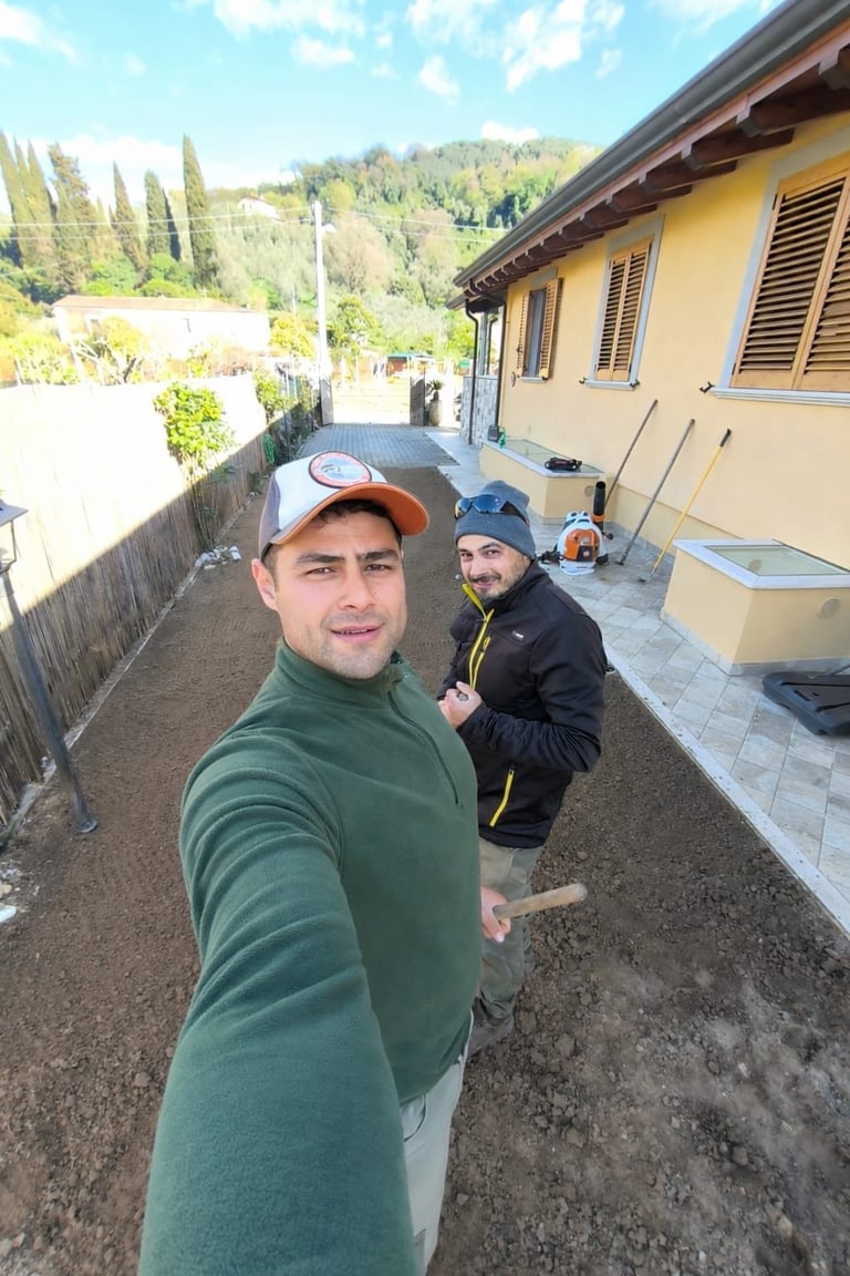 Two men smiling on a patio beside a yellow house with mountains and cypress trees in the background